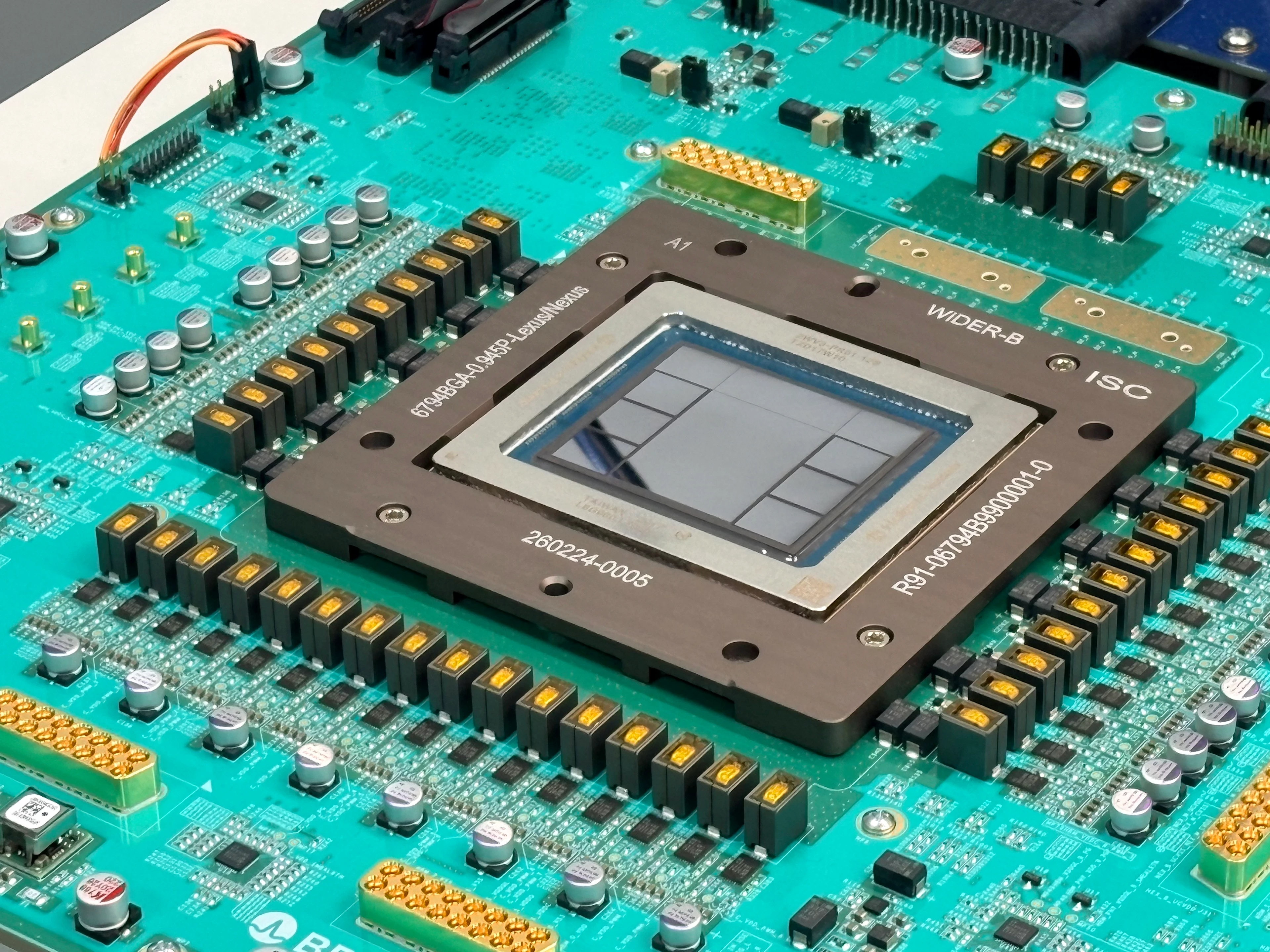 Nahaufnahme eines großen Prozessors mit freiliegendem Chip-Die auf einer türkisfarbenen Leiterplatte, umgeben von Kondensatoren, Steckverbindungen und weiteren elektronischen Bauteilen.